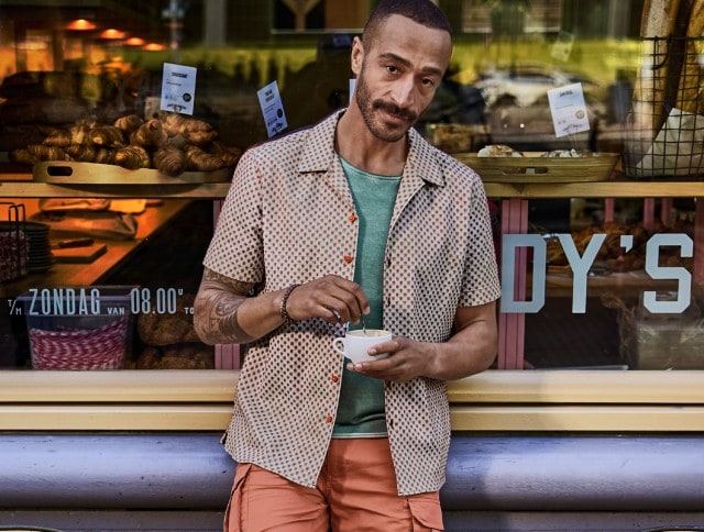 Ein Mann in einem gemustertem Hemd und einer roten Shorts steht vor einer Schaufensterscheibe und hat eine Tasse Kaffee in der Hand.