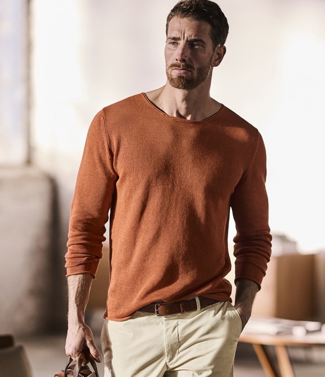 Ein Mann mir einem ziegelroten Pullover trägt eine Ledertasche und schaut entspannt zur Seite