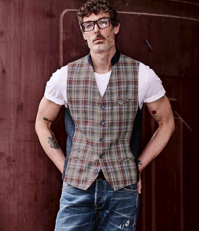 Ein Mann bekleidet mit einer Jeans und einer karierten Weste steht vor einer Wand und blickt frontal in die Kamera.