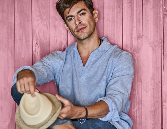 Ein Mann bekleidet mit einem hellblauen Leinenhemd und einer dunklen Jeans hält einen Sonnenhut in der Hand und sitzt vor einer Holzwand