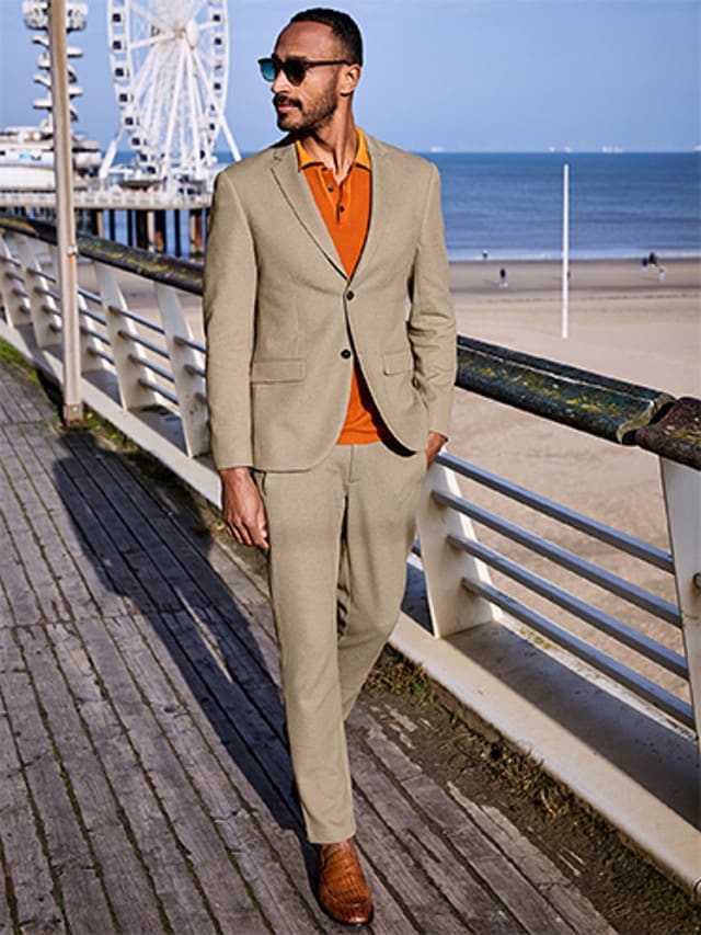 Ein Mann bekleidet mit einem hellbraunen Anzug und einem orangefarbenen Hemd läuft über eine Brücke am Strand entlang.