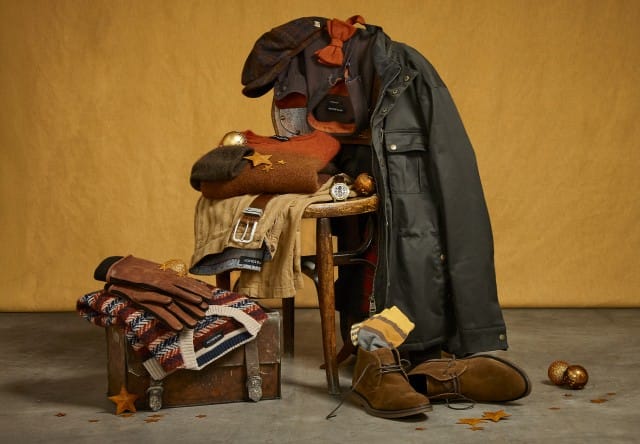 Das Bild zeigt ein warm zusammengestelltes Herrenoutfit mit Jacke, Pullover, Hose, Schuhen und Accessoires in Herbstfarben vor goldenem Hintergrund.