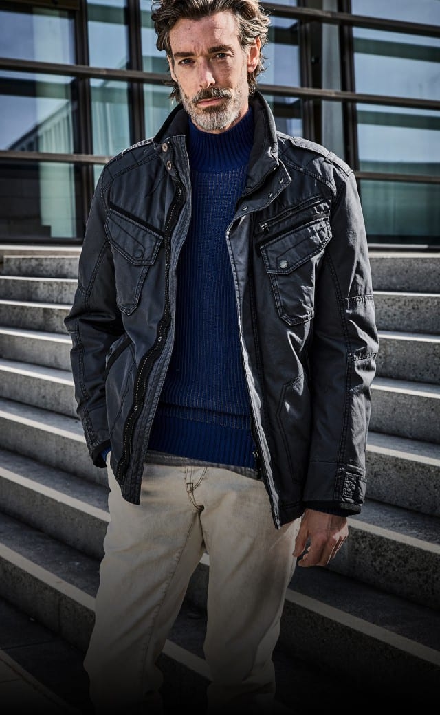 Ein Mann mit grauem Bart trägt eine dunkle Jacke über einem blauen Pullover und einer hellen Hose und steht auf einer Treppe vor einem modernen Gebäude. Ein Mann mit grauem Bart trägt eine dunkle Jacke über einem blauen Pullover und einer hellen Hose und steht auf einer Treppe vor einem modernen Gebäude.