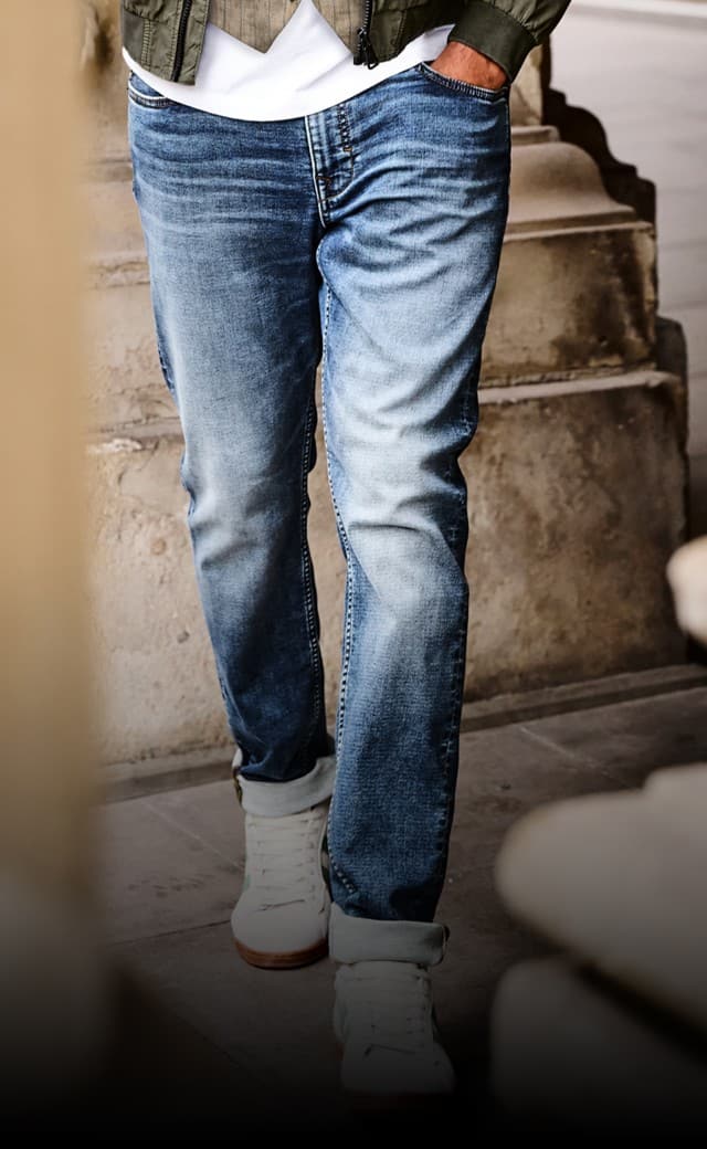 Ein Mann trägt eine verwaschene blaue Jeans mit hochgekrempelten Hosenbeinen, weiße Sneaker, ein weißes T-Shirt und eine grüne Jacke. Ein Mann trägt eine verwaschene blaue Jeans mit hochgekrempelten Hosenbeinen, weiße Sneaker, ein weißes T-Shirt und eine grüne Jacke.