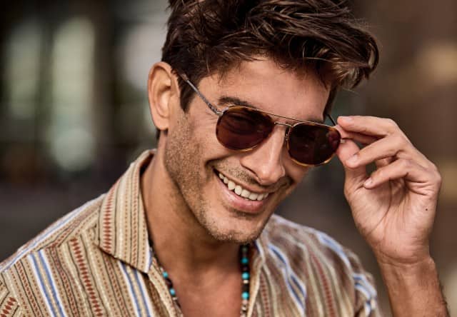 Eine Person trägt eine getönte Sonnenbrille, berührt diese leicht mit der Hand und ist nah aufgenommen vor einem unscharfen Hintergrund zu sehen. Eine Person trägt eine getönte Sonnenbrille, berührt diese leicht mit der Hand und ist nah aufgenommen vor einem unscharfen Hintergrund zu sehen.