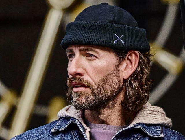 Der Mann trägt eine dunkelblaue Mütze, eine Jeansjacke mit hellem, flauschigem Innenfutter und darunter einen hellen Kapuzenpullover.