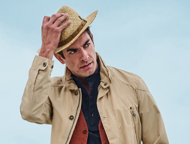 Auf dem Bild ist ein Mann zu sehen, der einen Strohhut, eine beige Jacke, eine rostrote Weste und ein dunkelblaues Hemd trägt.