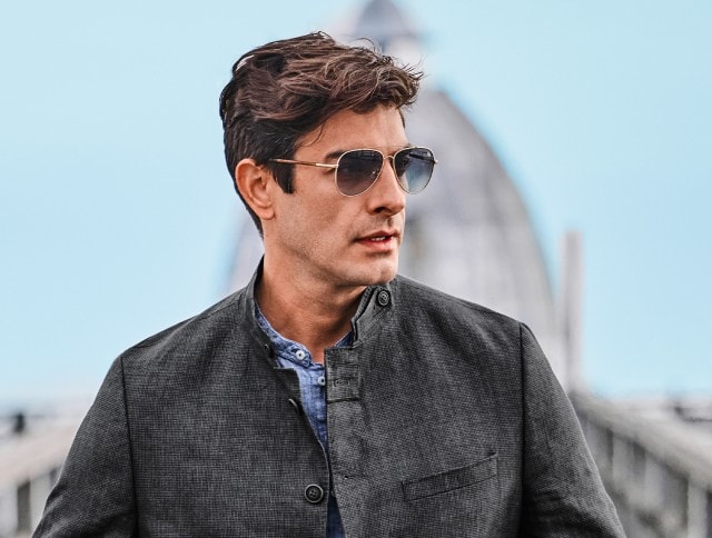Auf dem Bild ist ein Mann mit Sonnenbrille zu sehen, der eine graue, hochgeschlossene Jacke und ein blaues Hemd trägt.