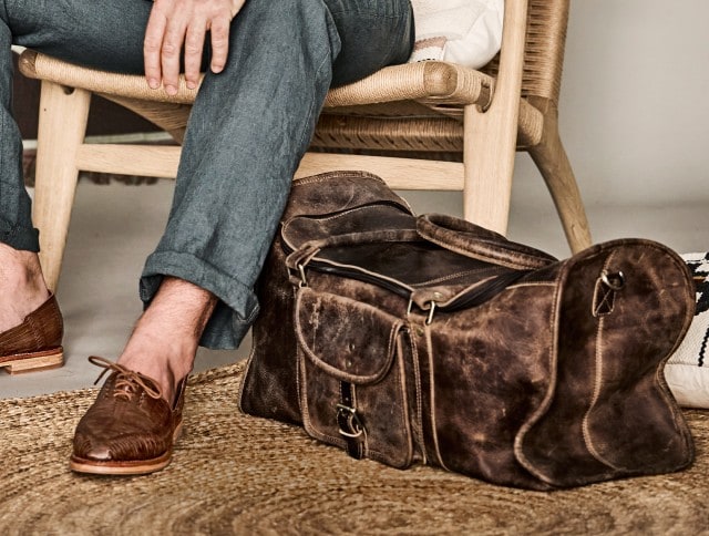 Auf dem Bild sitzt ein Mann in blauer Leinenhose und braunen Lederschuhen auf einem Stuhl, neben ihm steht eine große, braune Ledertasche auf dem Boden.