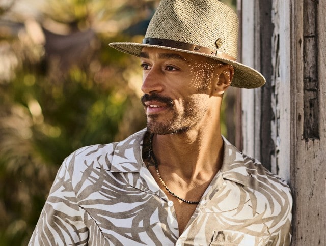 Ein Mann trägt ein beiges Sommerhemd mit weißem, abstraktem Muster und kombiniert dazu einen stilvollen Strohhut. Der Look wirkt lässig und zugleich elegant – perfekt für warme Tage und entspannte Stunden im Freien.