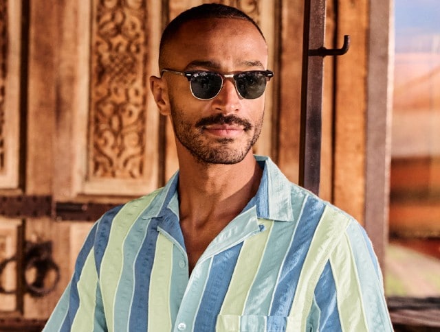 Ein Mann trägt ein sommerliches Hemd mit breiten, vertikalen Streifen in Hellblau, Grün und Gelb. Die dunkle Sonnenbrille rundet den entspannten, modischen Look perfekt für warme Tage ab.