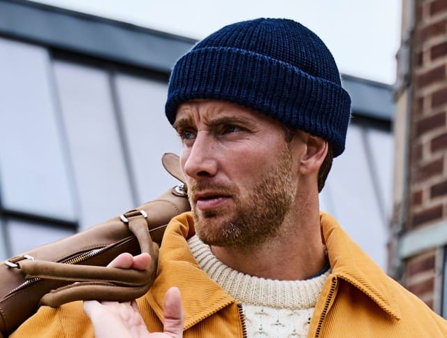 Der Mann trägt eine dunkelblaue Strickmütze zu einem cremefarbenen Zopfpullover und einer senfgelben Cordjacke – ein lässiger, farblich harmonischer Look mit herbstlichem Flair.