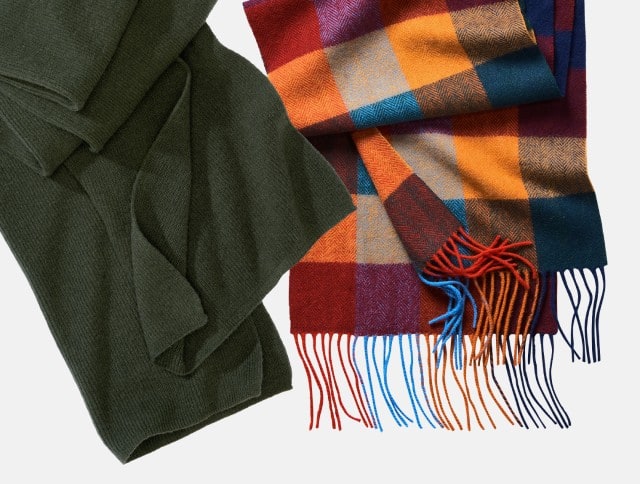 Zu sehen sind zwei Schals – einer in schlichtem Olivgrün aus fein gestricktem Material und der andere in einem farbenfrohen Karomuster aus Orange-, Rot-, Blau- und Gelbtönen mit Fransen – beide ideal für einen warmen, stilvollen Herbstlook.