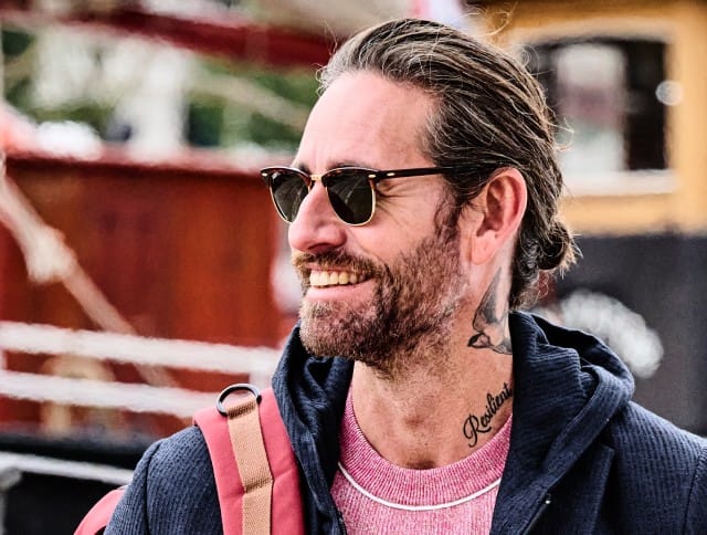 Ein Mann trägt eine dunkle Sonnenbrille, eine blaue Sweatshirtjacke und blickt lächelnd zur Seite.