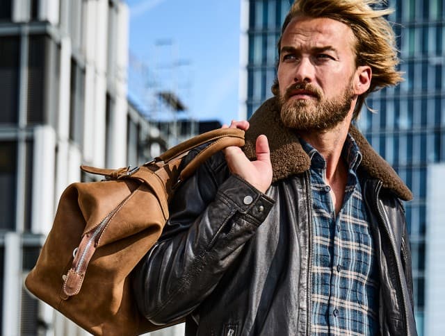 Der Mann trägt eine dunkelbraune Lederjacke mit Lammfellkragen über einem blau karierten Hemd und hat eine cognacfarbene Ledertasche lässig über der Schulter – ein markanter, maskuliner Look mit Vintage-Flair.