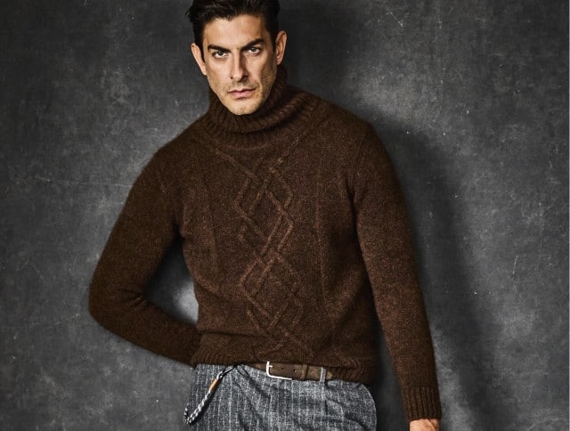 Ein Mann trägt einen braunen, grob gestrickten Rollkragenpullover mit Zopfmuster, kombiniert mit einer grauen, gemusterten Hose und braunem Gürtel.