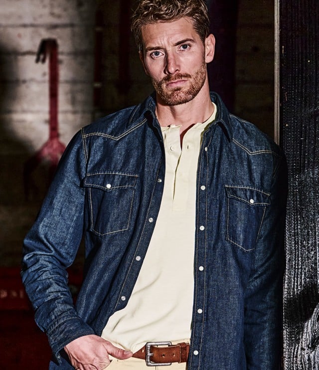 Ein Mann lehnt mit verschränkten Armen an einem Tisch und trägt ein beigebraun kariertes Hemd in einem Atelier‑ähnlichen Raum.