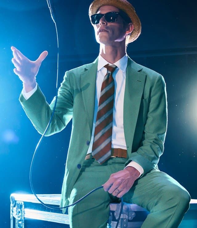 Ein Mann trägt einen hellgrünen Anzug mit weißem Hemd, braun‑grün gestreifter Krawatte, braunem Gürtel, Sonnenbrille und Strohhut.