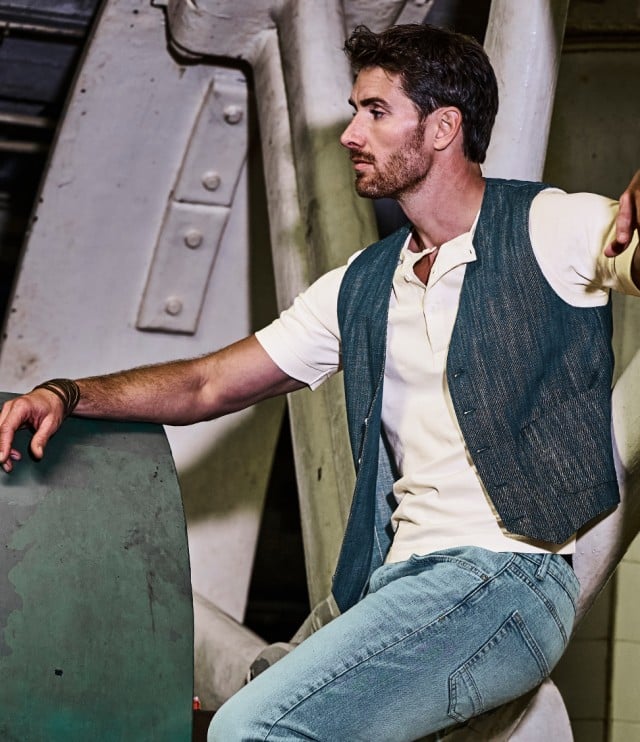 Ein Mann trägt ein cremefarbenes Poloshirt unter einer blau strukturierten Weste, kombiniert mit einer hellblauen Jeans.