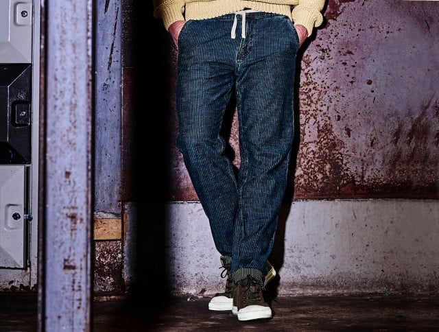 Auf dem Bild ist der Unterkörper eines Mannes zu sehen, der sich lässig vor einer Ziegelwand lehnt; er trägt blaue, karierte Stoffhosen und braune Wildlederstiefel.