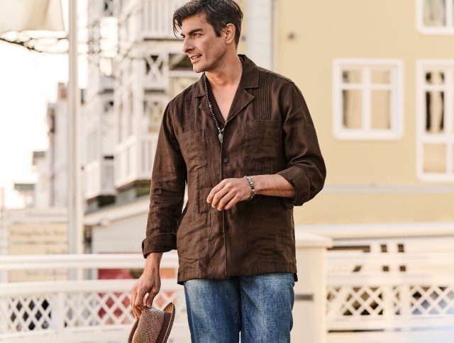 Auf dem Bild ist ein Mann zu sehen, der ein braunes langärmliges Leinenhemd und eine blaue Jeans trägt und einen Strohhut in der Hand hält.
