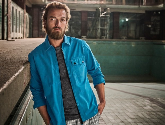 Auf dem Bild ist ein Mann zu sehen, der ein graues T-Shirt, ein offenes, hellblaues Hemd und karierte Shorts trägt.