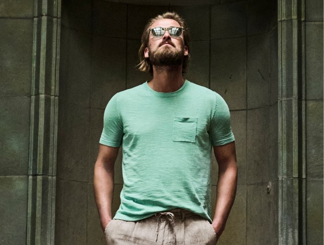 Auf dem Bild ist ein Mann mit Sonnenbrille zu sehen, der ein mintgrünes T-Shirt mit Brusttasche und eine hellbeige Leinenhose trägt.