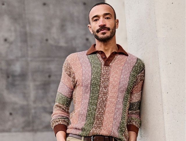 Auf dem Bild ist ein Mann zu sehen, der einen gestrickten Pullover mit vertikalen Streifen in Braun-, Grün- und Rosatönen sowie einen braunen Kragen trägt.