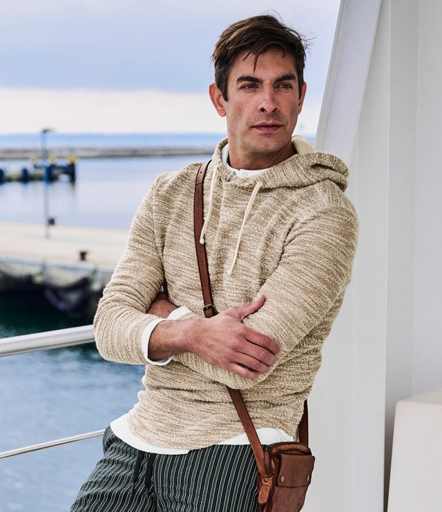 Auf dem Bild ist ein Mann zu sehen, der einen beigen Kapuzenpullover, eine grün-weiß gestreifte Shorts und eine braune Umhängetasche trägt.
