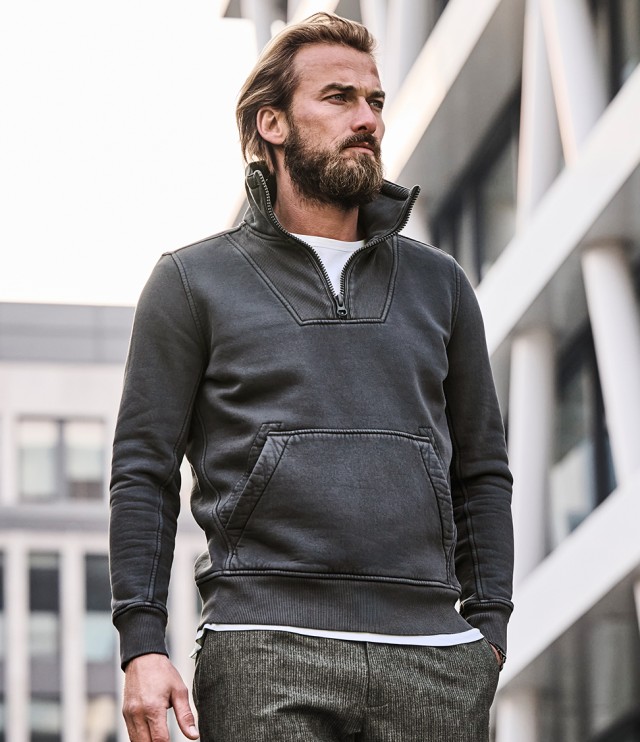 Auf dem Bild ist ein Mann zu sehen, der einen dunkelgrauen Pullover mit Reißverschluss am Kragen und Kängurutasche sowie eine graue Hose trägt.