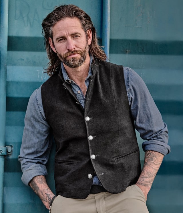 Auf dem Bild ist ein Mann zu sehen, der ein blaues Jeanshemd mit hochgekrempelten Ärmeln, eine schwarze Weste und eine beige Hose trägt.