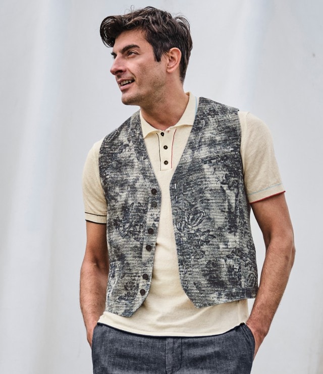 Auf dem Bild ist ein Mann zu sehen, der ein beigefarbenes Poloshirt mit Knopfleiste, eine gemusterte Weste und eine dunkelgraue Hose trägt.