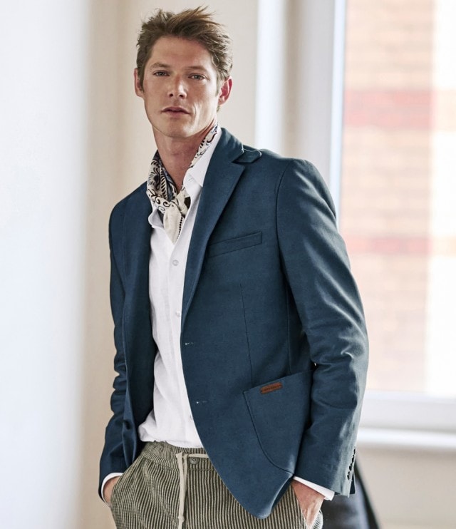 Auf dem Bild ist ein Mann zu sehen, der ein weißes Hemd, ein blaues Sakko, eine gemusterte Halstuch und eine gestreifte Hose trägt.