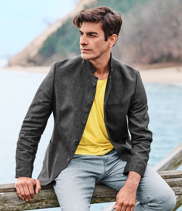 Auf dem Bild ist ein Mann zu sehen, der ein gelbes T-Shirt, eine graue, hochgeschlossene Jacke und eine hellblaue Jeans trägt.