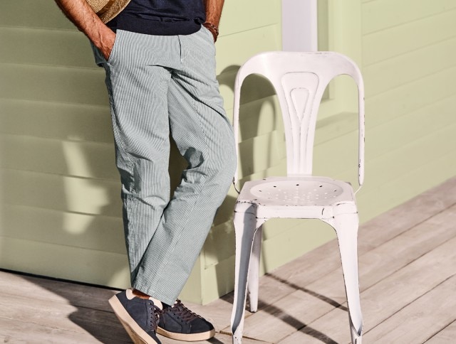 Auf dem Bild ist ein Mann zu sehen, der eine hellgrau-weiß gestreifte Hose, ein dunkelblaues Shirt und dunkelblaue Sneaker trägt; neben ihm steht ein weißer Stuhl.