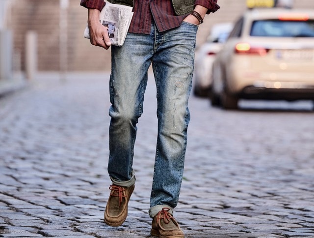 Auf dem Bild ist ein Mann zu sehen, der eine blau verwaschene Jeans, braune Wildlederschuhe und ein rot-braun kariertes Hemd trägt und eine Zeitung in der Hand hält.