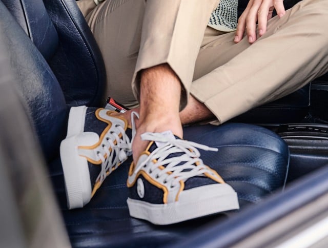 Auf dem Bild ist ein Mann zu sehen, der beigefarbene Hosen und dunkelblaue Sneaker mit weißen Sohlen und gelben Details trägt, während er auf einem blauen Ledersitz sitzt.