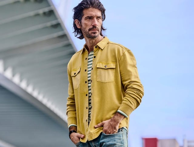 Ein Mann trägt ein senfgelbes Overshirt mit Brusttaschen über einem grün‑weiß gestreiften Shirt und kombiniert dazu eine hellblaue Jeans.