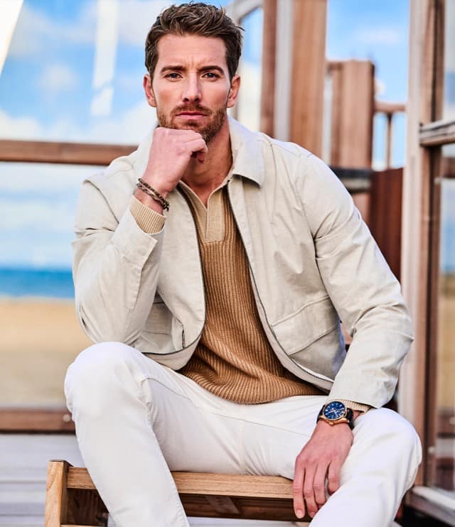 Ein Mann trägt eine hellbeige Jacke über einem beigefarbenen, gerippten Strickpullover mit Polokragen und dazu eine weiße Hose.