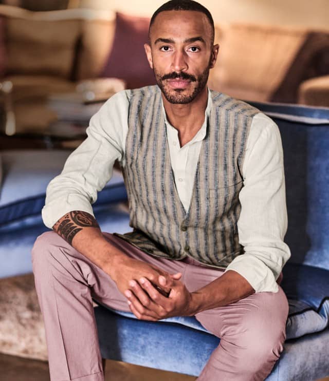 Ein Mann trägt ein helles, langärmliges Hemd unter einer gestreiften Weste und kombiniert dazu eine rosafarbene Hose.