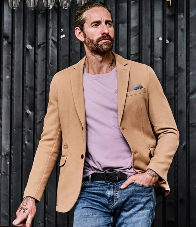 Ein Mann trägt ein beigefarbenes Sakko über einem hellvioletten T‑Shirt und kombiniert dazu eine blaue Jeans mit schwarzem Gürtel.