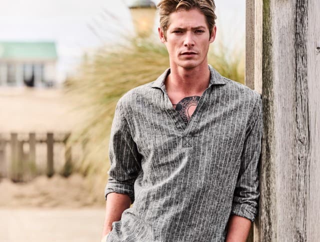 Ein Mann trägt ein grau gestreiftes Schlupfhemd und blickt ernst in die Kamera, hinter ihm ist ein Gebäude am Strand zu sehen