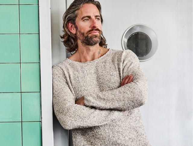 Ein Mann trägt einen hellen, pastellfarben melierten Strickpullover und lehnt lässig an einer blau gekachelten Wand.