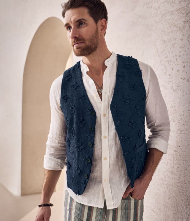 Ein Mann trägt ein helles, langärmliges Hemd unter einer gestreiften Weste und kombiniert dazu eine gestreifte Hose.