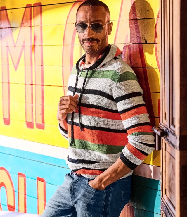 Ein Mann trägt einen lässigen Kapuzenpullover mit breiten, bunten Streifen in Weiß, Schwarz, Grün und Orange. Dazu kombiniert er eine blaue Jeans und Sonnenbrille – ein entspannter, moderner Freizeitlook für sonnige Tage.