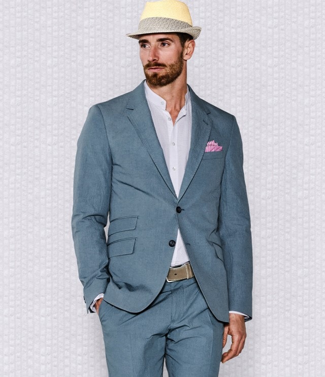 Ein Mann trägt einen hellblauen Sommeranzug mit weißem Hemd und beiger Kordel sowie einem hellen Strohhut. Das farbige Einstecktuch und der leichte Stoff verleihen dem eleganten Look eine entspannte, sommerliche Note.