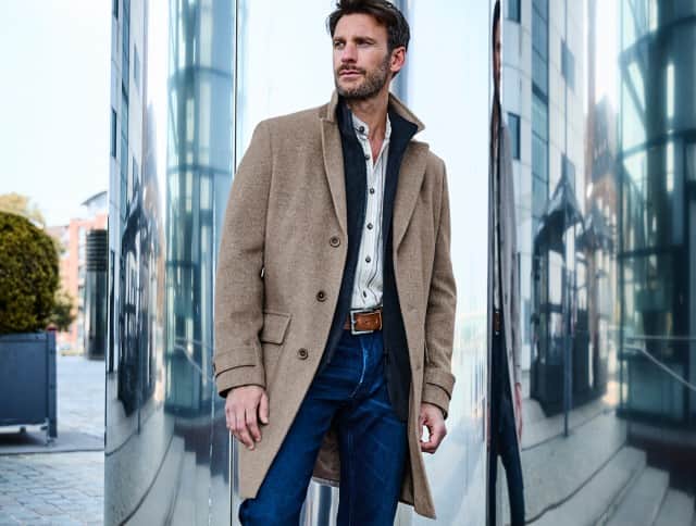 Der Mann trägt einen camel­farbenen Mantel über einer gestreiften Hemdjacke, kombiniert mit einer blauen Jeans und braunem Ledergürtel – ein moderner, zugleich klassischer Look mit urbaner Eleganz.