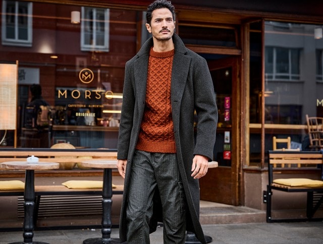 Ein Mann trägt einen langen dunklen Mantel über einem orangen Strickpullover und einer dunklen Hose, während er vor einem Café vorbeigeht.