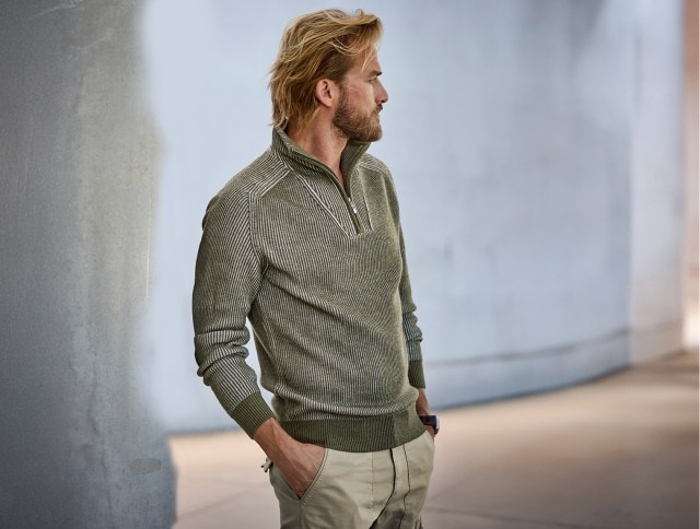 Ein Mann mit blondem Haar und Bart trägt einen olivgrünen Strickpullover mit Reißverschlusskragen und eine beige Hose, während er lässig mit den Händen in den Taschen zur Seite blickt.