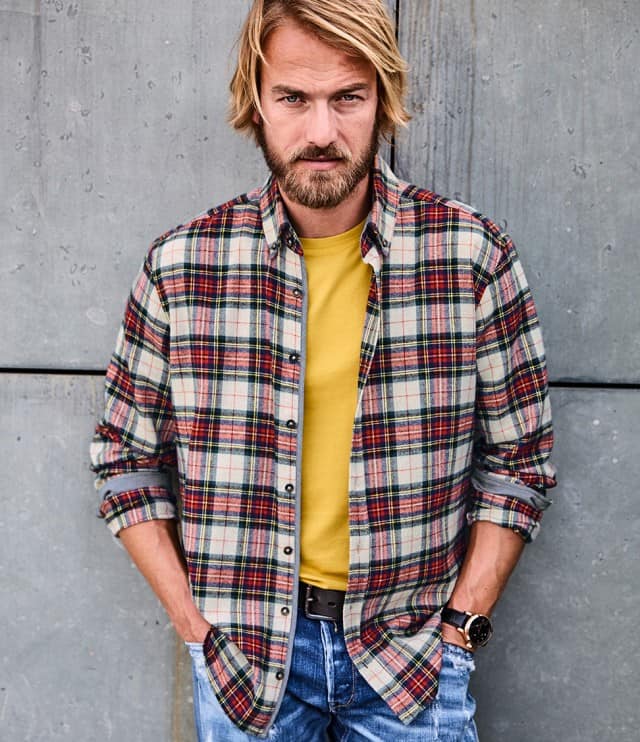 Der Mann trägt ein kariertes Flanellhemd in Rot-, Grün- und Beigetönen über einem senfgelben T-Shirt, kombiniert mit blauer Jeans – ein lässiger, farblich harmonischer Freizeitlook mit rustikalem Touch.